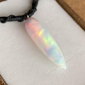 May include: A pear-shaped, iridescent opal pendant necklace. The pendant is white with flashes of pink, yellow, and green. It hangs from a black, woven cord necklace. The necklace is presented in a brown box with a white interior.