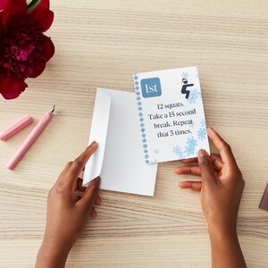 Könnte beinhalten: Eine blaue und weiße Karte mit Schneeflocken und dem Text "1st 12 squats. Take a 15 second break. Repeat that 3 times." Die Karte wird von den Händen einer Person gehalten.