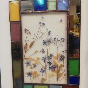 Puede incluir: Una obra de arte enmarcada en vitral con flores y hojas prensadas en tonos de azul, verde y amarillo. El marco está hecho de pequeños trozos de vidrio de varios colores, incluyendo rojo, azul, verde y amarillo.