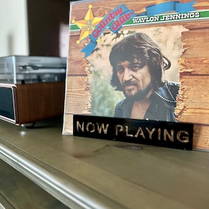 May include: A black sign with gold lettering that reads "NOW PLAYING" is in front of a vinyl record album cover. The album cover features a photo of Waylon Jennings and the text "Country  Guilt" and "Join the Country Club".
