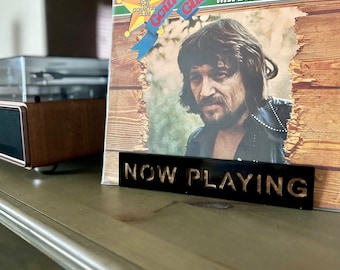 May include: A black sign with gold lettering that reads "NOW PLAYING" is in front of a vinyl record album cover. The album cover features a photo of Waylon Jennings and the text "Country  Guilt" and "Join the Country Club".