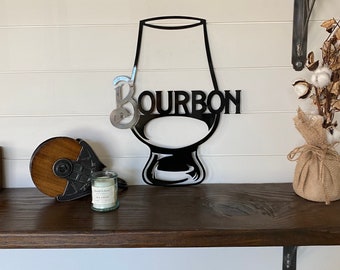 May include: Black metal wall decor shaped like a glass of bourbon with the word "BOURBON" written on it. The decor is mounted on a white wall with a wooden shelf above it.