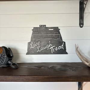 May include: Metal wall art featuring a silhouette of a truck with the words "Live Laugh Tread" written below. The art is mounted on a white wall with a wooden shelf below.