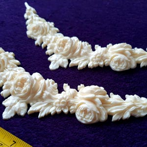 Pu&ograve; includere: Due applicazioni angolari floreali color crema con dettagli di rose e foglie. Le applicazioni sono disposte su uno sfondo viola. Un righello giallo nell'angolo in basso a sinistra misura circa 18 cm.
