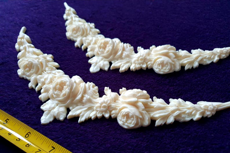 Pu&ograve; includere: Due applicazioni angolari floreali color crema con dettagli di rose e foglie. Le applicazioni sono disposte su uno sfondo viola. Un righello giallo nell'angolo in basso a sinistra misura circa 18 cm.