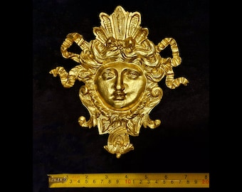 Può includere: Una scultura da parete dorata raffigurante un volto dettagliato con capelli fluenti, nastri e dettagli ornati. La scultura è lunga circa 20 cm, con un righello nell'immagine per la scala. Il pezzo è su sfondo scuro.