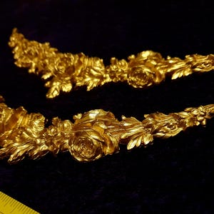 Pu&ograve; includere: Due ornamenti floreali dorati e ornati. Ogni pezzo presenta rose dettagliate, foglie e altri elementi floreali. Sono disposti su una superficie scura, con un righello giallo in primo piano, che suggerisce le dimensioni.