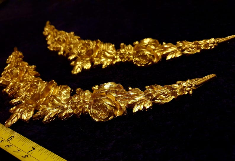 Pu&ograve; includere: Due ornamenti floreali dorati e ornati. Ogni pezzo presenta rose dettagliate, foglie e altri elementi floreali. Sono disposti su una superficie scura, con un righello giallo in primo piano, che suggerisce le dimensioni.