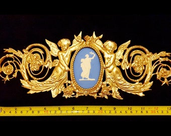 May include: An ornate gold wall decoration featuring two cherubs either side of an oval blue and white cameo. The cherubs are holding the cameo, which has a white silhouette. The decor is approximately 30 cm long.