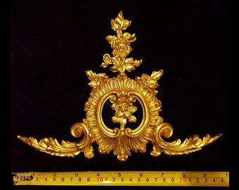 Può includere: Elemento architettonico decorativo dorato e ornato con motivi floreali e a volute. Il pezzo ha un design simmetrico con una forma ovale centrale. Il righello mostra una lunghezza di circa 23 cm.