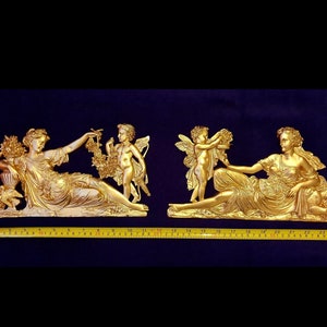 Può includere: Due sculture da parete color oro raffiguranti figure classiche. Una scultura mostra una donna reclinata con un cherubino che offre fiori. L'altra scultura mostra una donna reclinata con un cherubino. Le sculture sono lunghe circa 61 cm.