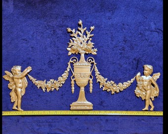 Può includere: Decorazione murale dorata con un'urna centrale con dettagli floreali, affiancata da due cherubini alati. Una ghirlanda floreale collega le figure e l'urna. L'opera è posta su uno sfondo blu, con un metro a nastro giallo in basso.