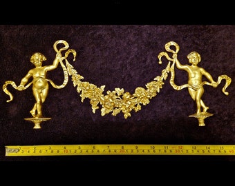 May include: Two gold cherub sculptures holding a floral garland with ribbons. The cherubs stand on decorative bases. A measuring tape shows the garland is approximately 40 cm long.