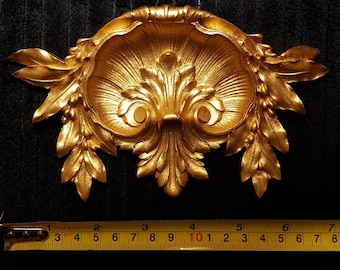 Può includere: Un'applique da parete decorativa dorata e ornata con un design simmetrico. L'applique presenta una forma centrale a conchiglia con dettagli di foglie e bacche. Il pezzo misura circa 18 cm di larghezza.