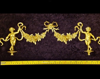 Può includere: Decorazione murale dorata con due figure di cherubini che tengono una ghirlanda floreale con un fiocco al centro. La ghirlanda è adornata con fiori e foglie. Il pezzo è lungo circa 46 cm.