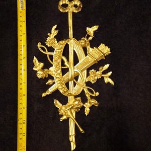 May include: A gold-colored metal wall ornament featuring a central oval frame, a quiver of arrows, and floral motifs, positioned next to a yellow measuring tape showing a total length of approximately 12 inches from top to bottom.