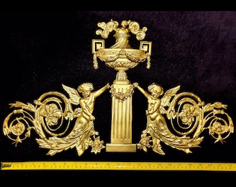 Può includere: Elemento architettonico dorato e ornato con una colonna centrale, un'urna decorativa e due cherubini alati. Il design include dettagli floreali e volute. Il pezzo è largo circa 61 cm.