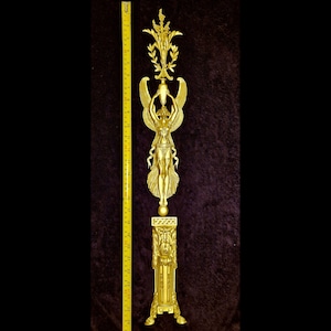 Peut inclure: Une sculpture décorative dorée représentant une figure ailée, des éléments floraux et une base en piédestal. La sculpture mesure environ 76 cm de haut, mesurée par un mètre ruban jaune. La pièce présente des détails complexes et un design classique.