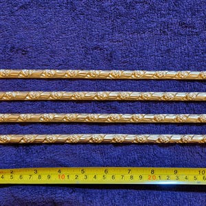 Könnte beinhalten: Vier goldfarbene, dekorative Metallstäbe mit floralem Design, parallel auf einer blauen Oberfläche angeordnet. Ein gelbes Maßband zeigt, dass die Stäbe etwa 30 cm lang sind.
