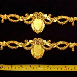 May include: Two ornate, gold-colored decorative appliques. Each features a central oval frame, a bow, and a garland of leaves. The appliques are approximately 10 inches long, ideal for furniture or craft projects.