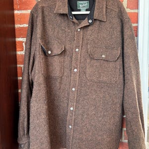 May include: A brown button-up shirt with two chest pockets and a black collar. The shirt is made of a textured fabric and is hanging on a white hanger. The shirt has silver buttons and a label on the collar.