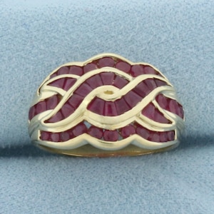 Puede incluir: Un anillo dorado con un diseño trenzado, adornado con numerosas piedras preciosas rojas oscuras de forma rectangular. Las piedras están engastadas muy juntas, creando un patrón continuo. El diseño intrincado del anillo es llamativo, adecuado para ocasiones especiales.