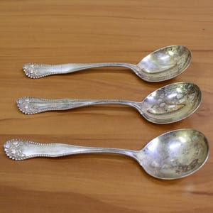 May include: Three silver spoons with ornate handles and a rounded bowl shape. The spoons are arranged in a row on a wooden surface.