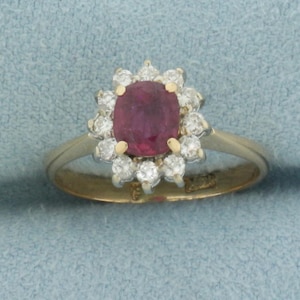 May include: A yellow gold ring featuring a central cushion-cut deep pinkish-red gemstone surrounded by a halo of twelve round brilliant-cut white diamonds. The setting uses gold prongs to secure the centre stone and the surrounding diamond cluster.
