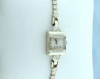 May include: A vintage wristwatch with a square face and diamond accents. The watch has a white face with black Roman numeral hour markers and a black hand. The watch is set in a gold-toned metal case and has a gold-toned metal band.