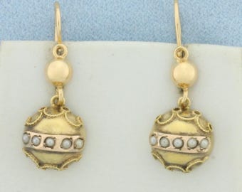 May include: A pair of gold-toned drop earrings. Each earring features a small gold ball at the top, connected to a larger, round, ornate gold pendant. The pendants have a band of small, white pearls. The earrings are on a white surface.