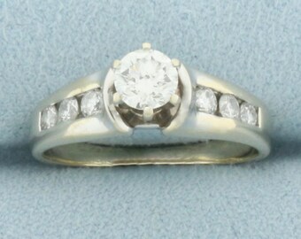 May include: A white gold ring featuring a large, round, clear diamond center stone. The band has a channel set of smaller diamonds on each side. The ring is set against a light blue background.