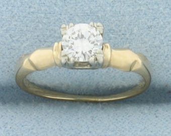 May include: A gold engagement ring with a large, round, clear diamond center stone. The ring has a classic design with a gold band and a four-prong setting. The ring is set against a light blue background.
