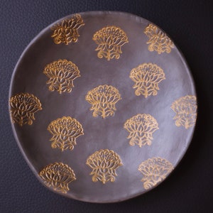 May include: A round, grey ceramic plate with a decorative pattern of gold floral designs.
