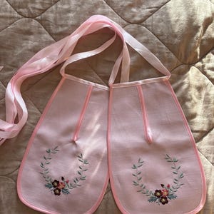 May include: Two light pink fabric bibs featuring a satin ribbon trim and long matching ties. Each bib is decorated with an embroidered wreath of green leaves and small dark red and pink flowers centered at the bottom of the fabric panel.
