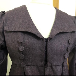 May include: A plum-colored, double-breasted jacket with a wide collar and large, round buttons. The jacket has a tailored fit and features a subtle herringbone pattern in the fabric. The sleeves are slightly puffed at the shoulders, adding a vintage touch.