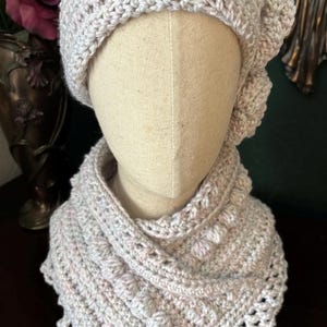 May include: A light gray crochet hat and cowl set. The hat has a flower detail on the side. The cowl has a textured, bobble stitch design. The set is displayed on a mannequin head.