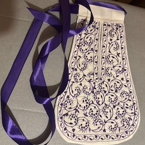 Könnte beinhalten: Ein weißer Stoffbeutel mit einem violetten Band. Der Beutel ist mit einer violetten Stickerei eines Blumendesigns verziert.