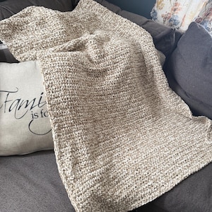 May include: A beige crocheted blanket draped over a gray sofa. The blanket has a thick, textured appearance, with visible stitches creating a cozy, handmade look. A decorative pillow with the words "Family is forever" is visible.