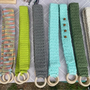 May include: Seven crocheted belts with wooden rings. The belts are in various colors, including green, blue, gray, white, and pink. The wooden rings are all the same size and are attached to the ends of the belts.