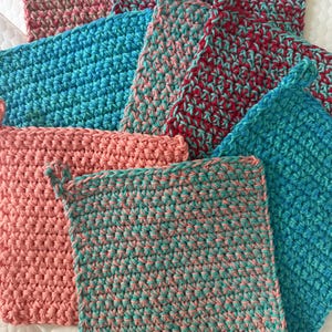 May include: A collection of handmade crochet potholders in various colors. The potholders are square-shaped with a loop for hanging. Colors include shades of pink, blue, and red, some with a speckled effect. The texture is dense and the stitches are clearly visible.