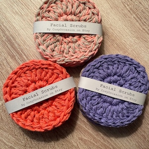 May include: Three round, crocheted facial scrub pads in coral, lavender, and a pink and gray mix. Each pad has a white paper band with the text "Facial Scrubs By CozyOccasion on Etsy". The pads are arranged on a light brown wooden surface.