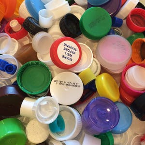 May include: A collection of colorful plastic bottle caps, some with text, including "SHAKE WELL BEFORE OPENING" and "BESTBY 11/17/16 T1 1:1751-0500058839".