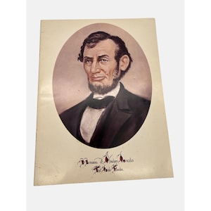 May include: A vintage, cream-colored card with a portrait of Abraham Lincoln. The portrait is in a circular frame and shows Lincoln in a dark suit with a white shirt and a black bow tie. The text "Memories of Abraham Lincoln" is written in red cursive at the bottom of the card.
