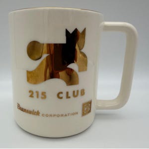 May include: A white ceramic mug featuring a large, reflective gold graphic design. The text 215 CLUB is printed in gold below the graphic, with Brunswick CORPORATION and a small square logo containing the letter B printed in gold at the bottom.