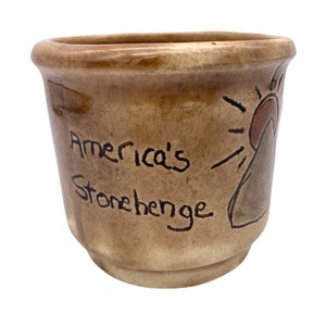 May include: A brown ceramic mug with the words "America's Stonehenge" written on it. The mug has a brown and orange sun design on the side.