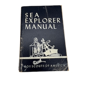 以下が含まれることがあります： ボーイスカウト・オブ・アメリカのヴィンテージの黒いSea Explorer Manual。表紙には白い文字と、船のランタン、双眼鏡、六分儀などの航海用具のイラストが描かれています。本には使用感が見られます。
