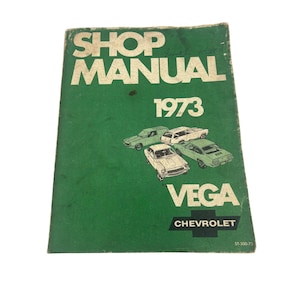 May include: A vintage shop manual for the 1973 Chevrolet Vega. The cover is green with white text that reads "SHOP MANUAL 1973 VEGA CHEVROLET." Illustrations of cars are featured on the front. The manual shows signs of wear.