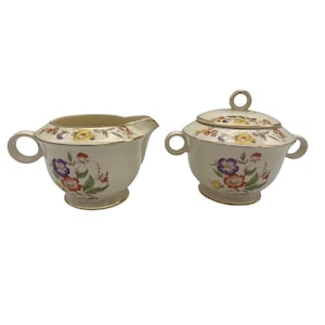 May include: Cream-colored creamer and sugar bowl set with floral designs. The creamer has a handle and spout, while the sugar bowl has a lid with a loop handle. Both pieces feature colorful flowers and gold trim, creating a vintage aesthetic.