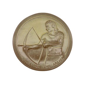 Puede incluir: Medallón redondo de color bronce con un relieve de un hombre disparando un arco y una flecha. El texto "JONATHAN THE ARCHER" está inscrito en el borde inferior. El arquero lleva una camiseta sin mangas.