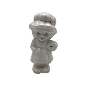 May include: A white porcelain figurine of a girl wearing a white hat with gold trim and a white dress with gold trim. She is holding a basket in her right hand.